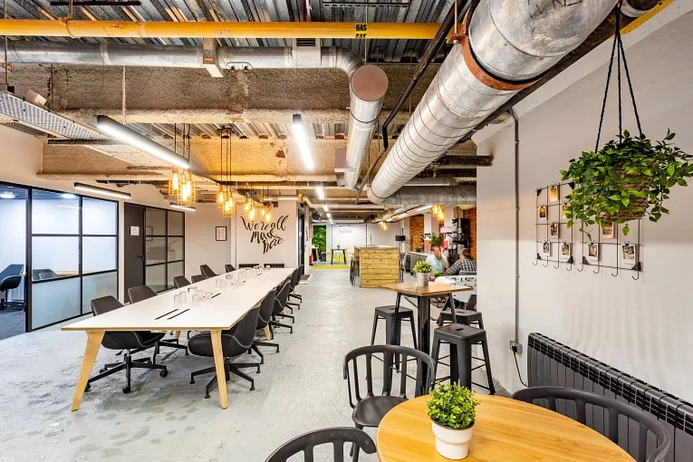 Open plan coworking space in Exeter featuring communal tables, exposed metal pipe ceilings, indoor plants, and modern seating areas.