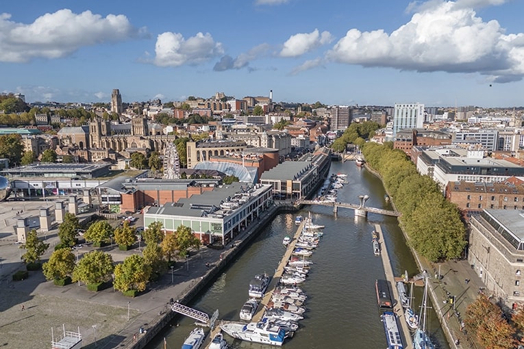 Aerial view of Bristol Harbourside with boats, commercial properties, and city skyline under a blue sky with clouds.