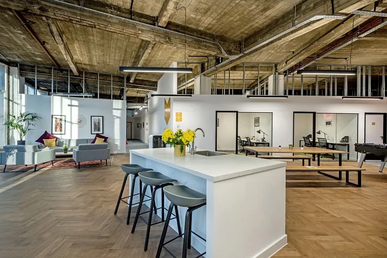 Open-plan coworking space with a kitchen island, lounge seating, herringbone floors, and glass-front private offices flooded with natural light.