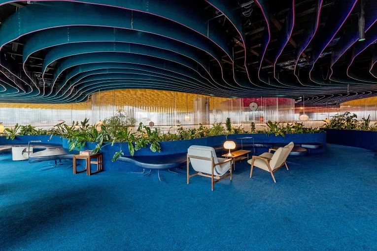 Modern coworking space lounge with blue sculptural ceiling, plants, and mixed seating in London.