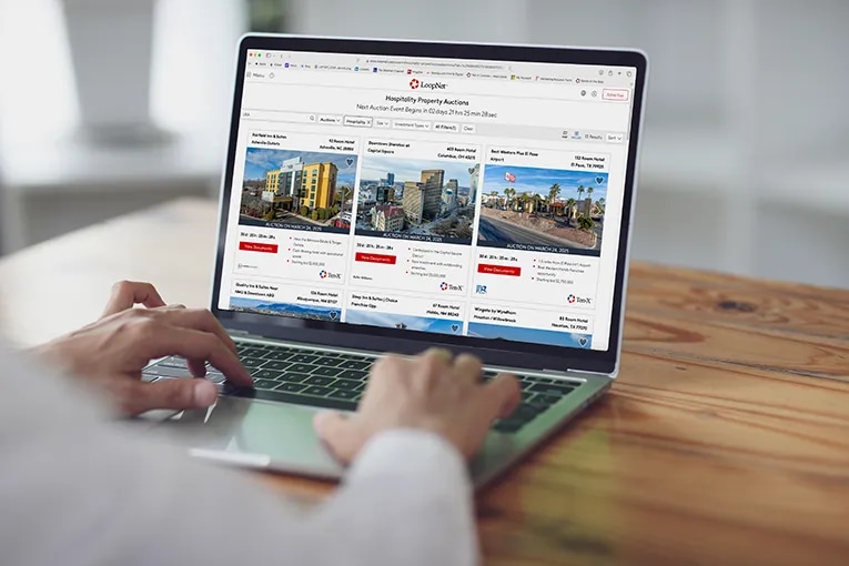 Person using a laptop displaying LoopNet's hospitality property auctions page. The screen shows three commercial property listings with images of hotels/buildings and auction details. The user's hands are visible typing on the keyboard as they browse available auction properties on the wooden desk.
