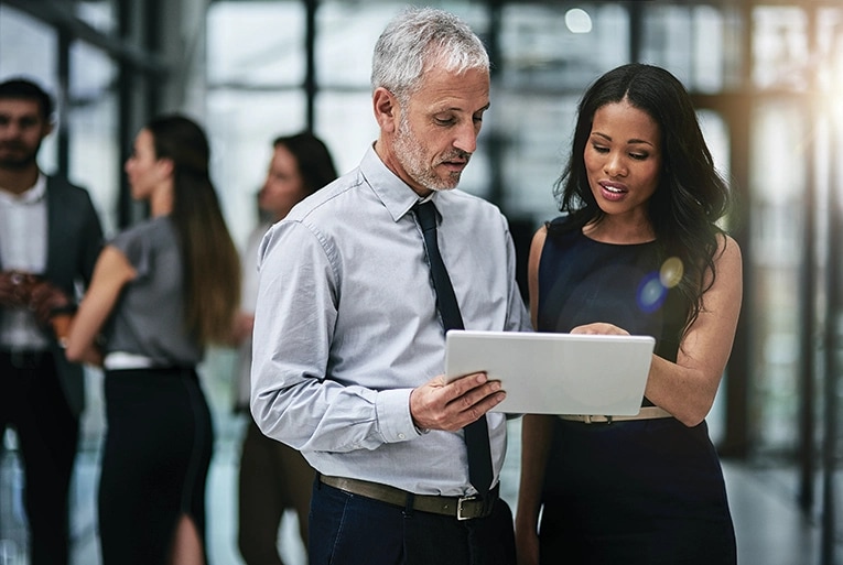 Two business professionals reviewing auction information on a laptop in a modern office environment. A well-dressed man with gray hair and a woman in a black dress are collaborating while examining property details, with other colleagues visible in the background. This represents the professional nature of commercial real estate auctions conducted through digital platforms.