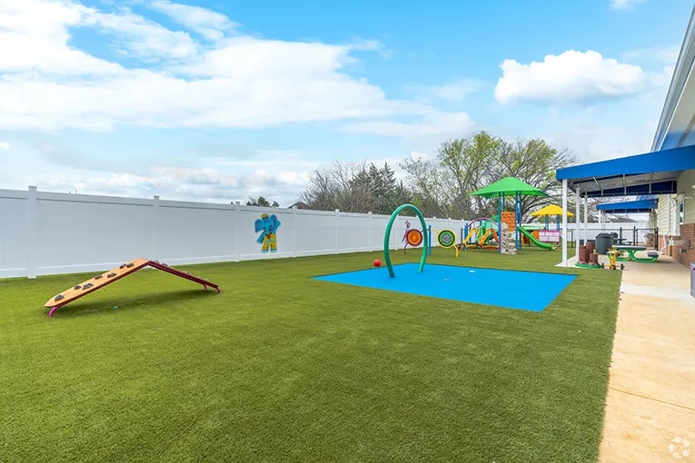 Exterior view of The Learning Experience showing the outside area enclosed with a white wall, grass and playground equipment
