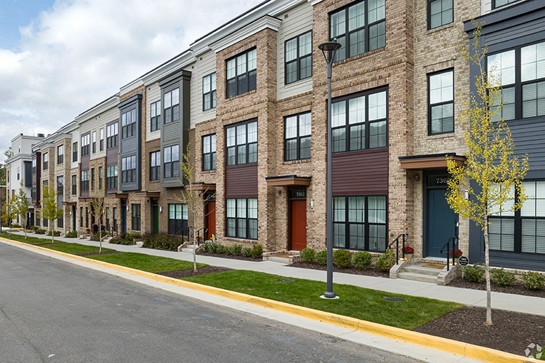 Row of modern three-story brick and siding apartment townhomes with individual front doors, sidewalk, young trees, and a streetlight.
