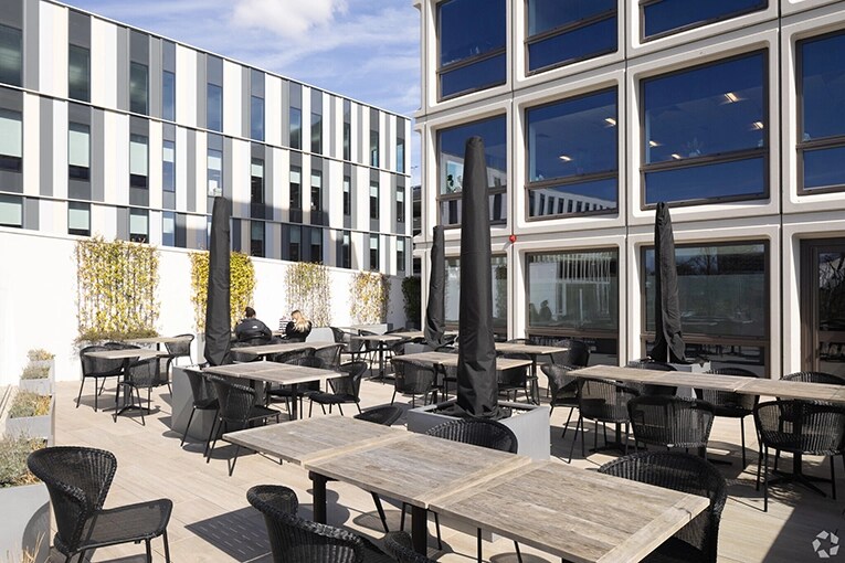Outdoor terrace of a coworking space in Hampshire, UK featuring multiple wooden tables with black wicker chairs. The space has a modern deck floor and is surrounded by contemporary architecture with striking geometric façades and large glass windows that reflect the blue sky. Several closed patio umbrellas in dark covers are placed throughout the area. Plantings add greenery along one wall, creating a pleasant outdoor work and meeting environment that complements the sleek commercial buildings.RetryClaude can make mistakes. Please double-check responses.