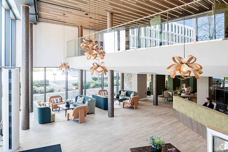 Modern coworking space in Hampshire, UK featuring a bright open lobby with high ceilings and wooden slat details. The space has a mezzanine level with glass railings, distinctive curved wooden pendant lights, and comfortable seating areas with blue and tan furniture. Large windows provide natural light, and there's a reception desk visible in the foreground. The contemporary design combines wood, glass, and natural elements creating a welcoming professional environment.
