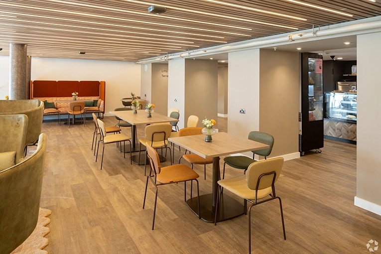 Modern café area within a coworking space in Hampshire, UK featuring wooden slatted ceiling and warm wood flooring. The space includes several small tables with mixed seating in green, beige, and orange chairs. A cozy booth with terracotta upholstery sits against the wall, and there's a small café counter visible in the background. Fresh flowers in small vases decorate each table, creating a welcoming atmosphere for casual meetings and work breaks.